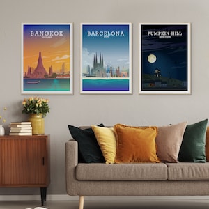 May include: Three framed city skyline posters with a minimalist design. The posters are hanging on a light gray wall above a gray couch with pillows. The posters feature the city skylines of Bangkok, Thailand, Barcelona, Spain, and Pumpkin Hill, United States.