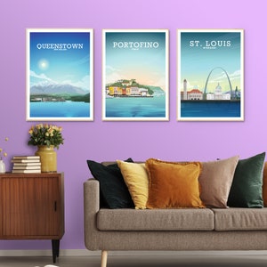 May include: Three framed travel posters with illustrations of Queenstown, New Zealand, Portofino, Italy, and St. Louis, Missouri. The posters are hanging on a light purple wall above a gray couch with pillows.