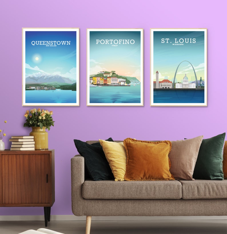 May include: Three framed travel posters with illustrations of Queenstown, New Zealand, Portofino, Italy, and St. Louis, Missouri. The posters are hanging on a light purple wall above a gray couch with pillows.