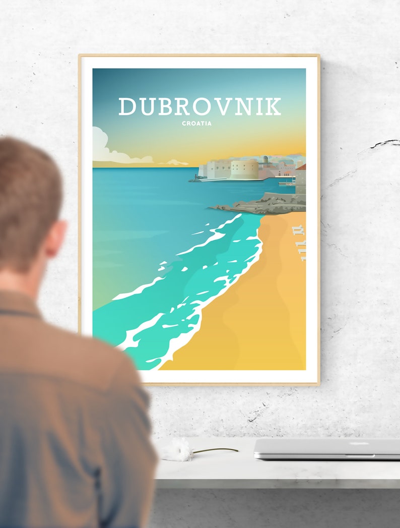 May include: A framed travel poster featuring the word "DUBROVNIK" above "CROATIA". The poster depicts a coastal scene with a turquoise sea, sandy beach, and a city with white buildings. The sky transitions from blue to yellow.