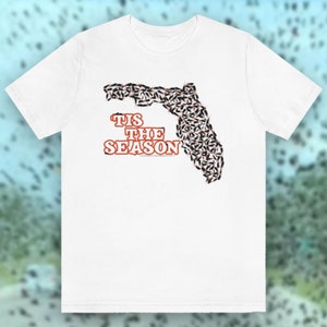 May include: White t-shirt with a graphic of the state of Florida made up of small black and orange insects. The text "'Tis the Season" is printed in orange below the graphic.