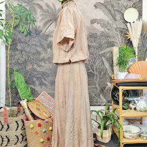 Op de afbeelding: Een beige maxi-jurk met een bruin stippenpatroon. De jurk heeft korte mouwen en een getailleerd bovenlijf. De jurk is op een mannequin tegen een achtergrond van een vintage botanische print.