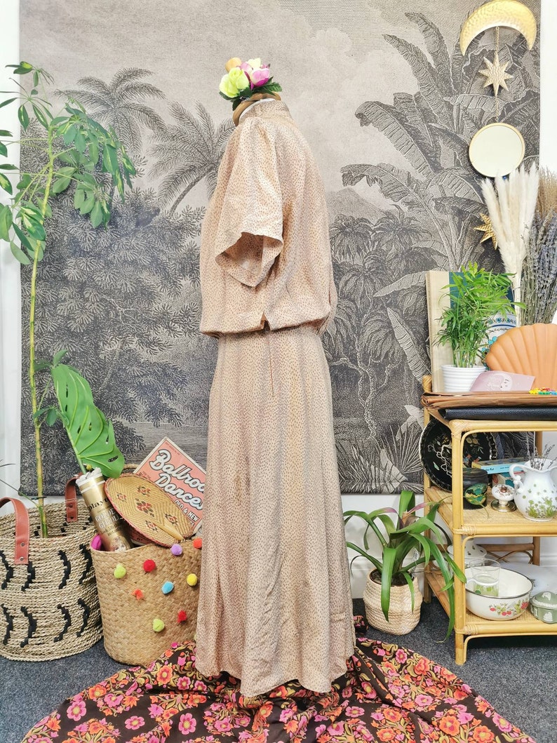 Op de afbeelding: Een beige maxi-jurk met een bruin stippenpatroon. De jurk heeft korte mouwen en een getailleerd bovenlijf. De jurk is op een mannequin tegen een achtergrond van een vintage botanische print.