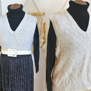 Könnte beinhalten: Eine weiße, gestrickte, ärmellose Strickweste mit V-Ausschnitt. Die Weste wird über einem schwarzen Rollkragenpullover und einem schwarzen Faltenrock mit einem weißen Gürtel getragen.