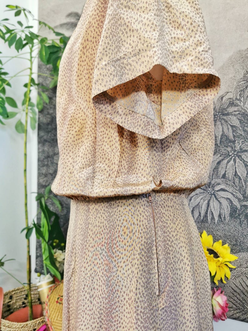 Op de afbeelding: Een beige jurk met een bruin stippenpatroon. De jurk heeft korte mouwen en een rits aan de achterkant.