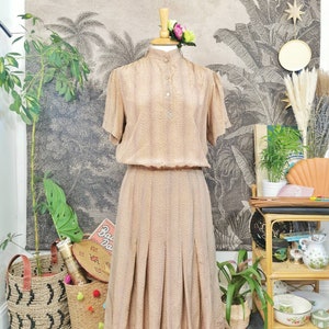 Op de afbeelding: Een beige jurk met een bruin stippenpatroon. De jurk heeft een hoge hals, korte mouwen en een geplooide rok. De jurk is op een mannequin tegen een achtergrond met een tropische print.