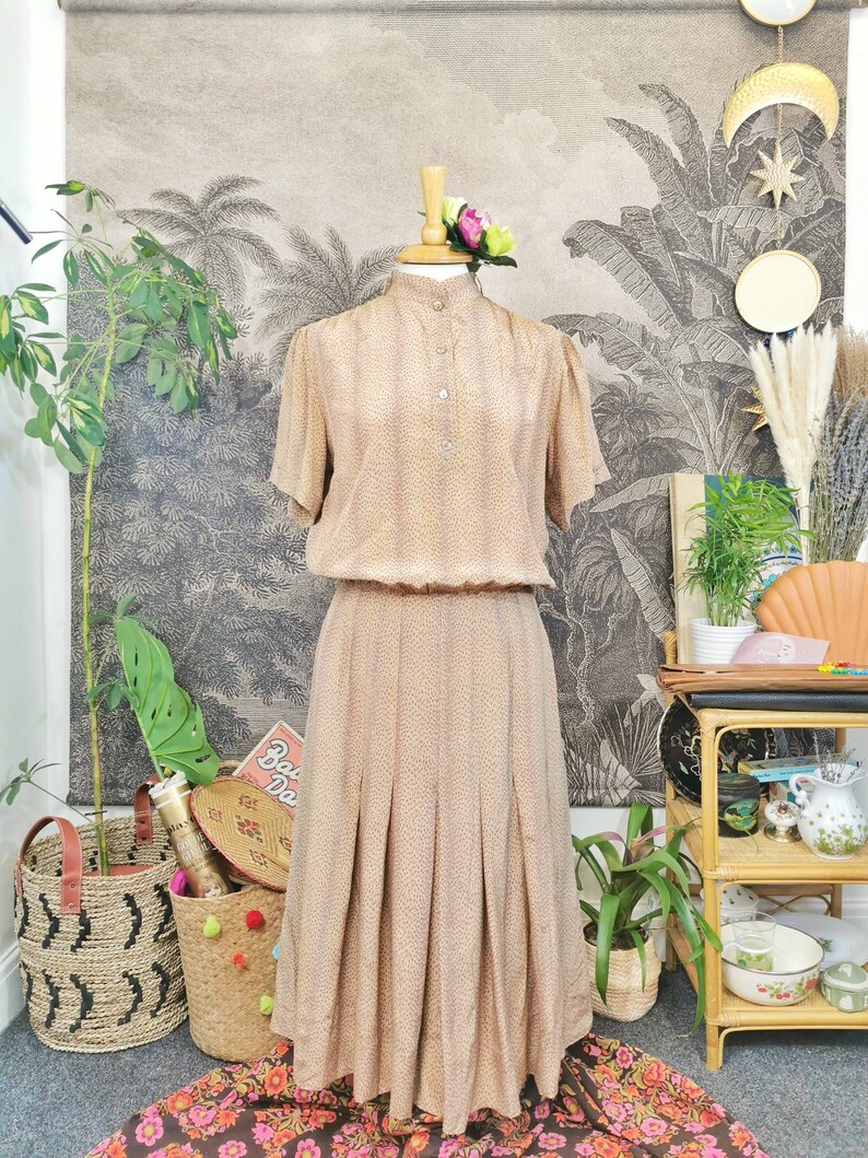 Op de afbeelding: Een beige jurk met een bruin stippenpatroon. De jurk heeft een hoge hals, korte mouwen en een geplooide rok. De jurk is op een mannequin tegen een achtergrond met een tropische print.