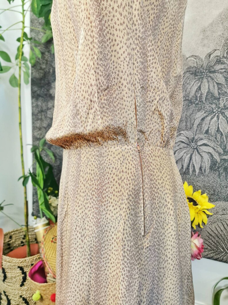 Op de afbeelding: Een beige jurk met een bruin gevlekt patroon. De jurk heeft een rits aan de achterkant en een getailleerde pasvorm.
