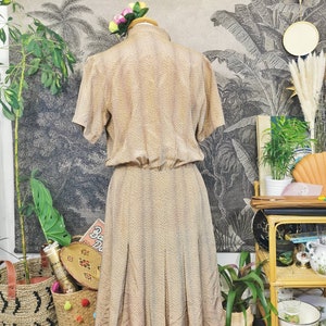 Op de afbeelding: Een beige midi-jurk met een klein bruin stippenpatroon. De jurk heeft korte mouwen en een aansluitend bovenlijf. De rok is geplooid en valt tot halverwege de kuiten.