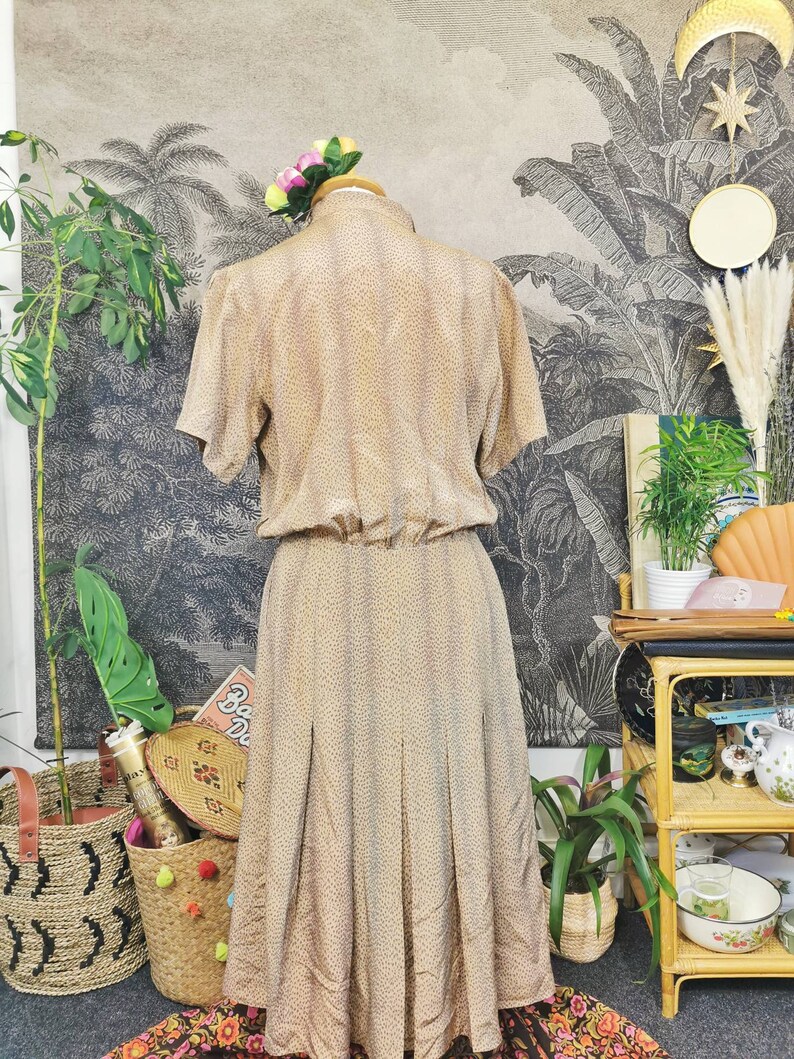 Op de afbeelding: Een beige midi-jurk met een klein bruin stippenpatroon. De jurk heeft korte mouwen en een aansluitend bovenlijf. De rok is geplooid en valt tot halverwege de kuiten.
