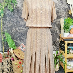 Op de afbeelding: Een vintage jurk in bruin en beige met een hoge kraag en een knoopsluiting aan de voorkant. De jurk heeft een getailleerd bovenstuk en een zwierige rok met plooien. De jurk is gemaakt van een lichtgewicht stof met een subtiel patroon.