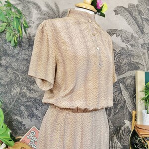 Op de afbeelding: Een beige jurk met kleine roze stippen. De jurk heeft een hoge kraag, korte mouwen en een knoopsluiting aan de voorkant. De jurk is op een mannequin tegen een achtergrond met een tropische print.