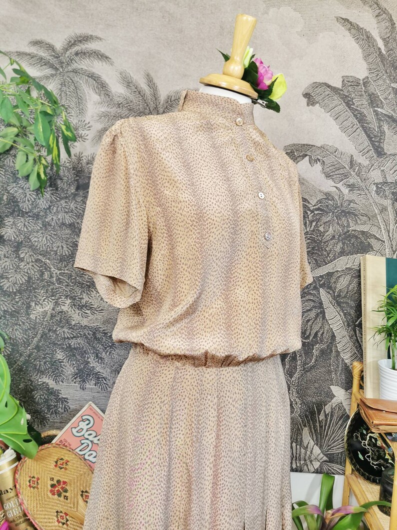 Op de afbeelding: Een beige jurk met kleine roze stippen. De jurk heeft een hoge kraag, korte mouwen en een knoopsluiting aan de voorkant. De jurk is op een mannequin tegen een achtergrond met een tropische print.
