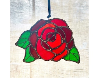 May include: Stained glass rose with red, burgundy, and orange petals, and green leaves. The floral design is outlined in a dark brown, with a black ribbon for hanging. The artwork is a decorative piece.