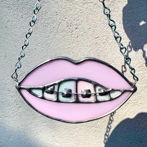May include: A stained glass piece depicting a pair of pink lips with braces. The lips are outlined in a dark metal, and the braces are silver. A silver chain is attached for hanging.