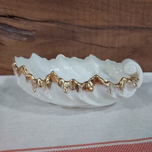 May include: A white ceramic dish shaped like a seashell with a gold rim. The dish is on a white and red striped fabric.
