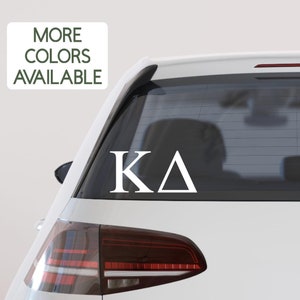 May include: White vinyl decal with the Greek letters "KA" on a white car. The decal is on the back window of the car.
