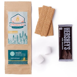 May include: A brown paper bag with a label that says "S'mores Kit by Eventful Sweets" and a smaller label that says "Eventful Sweets". The bag is next to two graham crackers, two white marshmallows, and a Hershey's milk chocolate bar.
