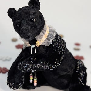Peut inclure: Un ours en peluche noir avec un col à volants et des ornements perlés. L'ours a un visage détaillé et est orné de petites boucles d'oreilles perlées colorées. Le jouet en peluche est posé sur un fond neutre et doux.