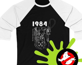 Op de afbeelding: Een zwart-wit langarm baseballshirt met een witte raglanmouw. Het shirt heeft een zwart-witte illustratie van een protonpack uit de film Ghostbusters met de tekst "1984" en "The Proton Pack is not a toy."  Een groene slijmsplatter bevindt zich in de rechterbenedenhoek met een witte geest met een rode cirkel en een schuine streep.