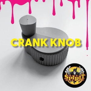 Puede incluir: Pomo de manivela de plástico gris con una superficie texturizada y una parte superior redonda. El pomo tiene las palabras "CRANK KNOB" impresas en amarillo en la parte delantera. El fondo es blanco con pintura rosa goteando por los lados.