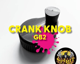 Puede incluir: Un pomo de manivela negro impreso en 3D con el texto "GB2" impreso en él. El pomo está sobre una superficie blanca con un diseño de pintura rosa salpicada.