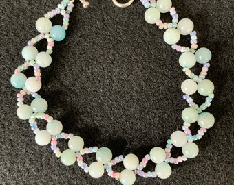 May include: A beaded bracelet featuring pale green and white round beads. The bracelet is connected with a silver toggle clasp. The design incorporates smaller, colorful seed beads in shades of pink, blue, and green, creating a delicate, handcrafted look.
