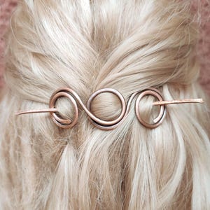 May include: Copper hair pin with an infinity symbol design. The pin is inserted into blonde hair, showcasing the intricate metalwork. The pin's design includes a series of loops and a straight bar for securing the hair.