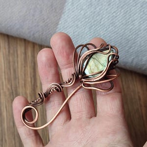 May include: A copper wire-wrapped brooch featuring a central marquise-shaped labradorite gemstone with iridescent green and gold flashes, surrounded by intricate swirling metal loops and small decorative beads.