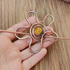 Puede incluir: Una horquilla para el pelo de alambre de cobre con un diseño de flor y una piedra preciosa amarilla en el centro.