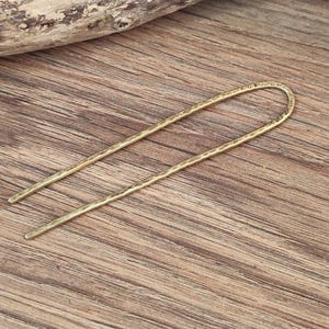 May include: A gold-toned hair stick with a hammered texture. The stick is long and thin, with a slight curve at the top.