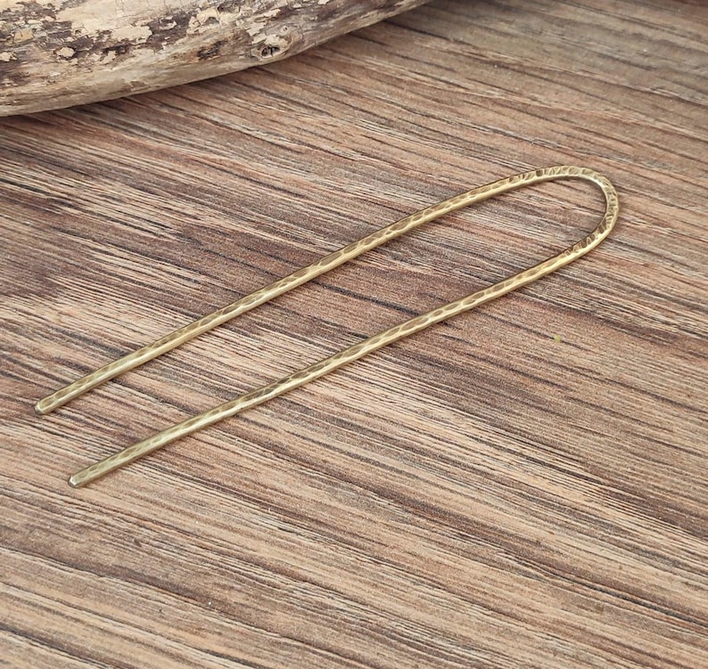 May include: A gold-toned hair stick with a hammered texture. The stick is long and thin, with a slight curve at the top.
