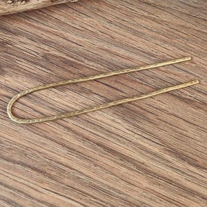 May include: A gold-toned metal hair stick with a hammered texture. The stick is bent into a U-shape.