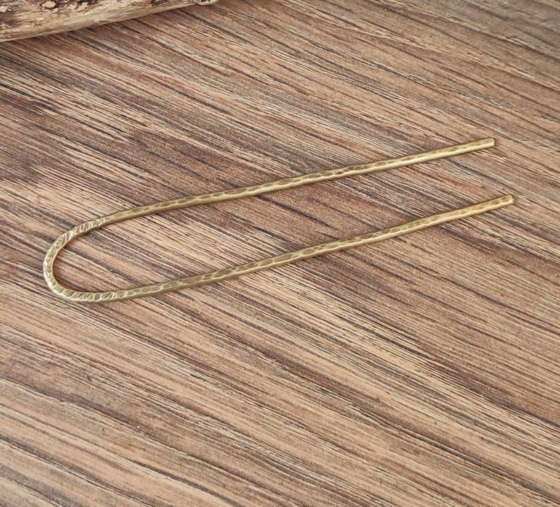 May include: A gold-toned metal hair stick with a hammered texture. The stick is bent into a U-shape.