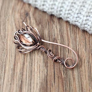 Könnte beinhalten: Eine kupferfarbene Haarspange aus Metall in Form einer Blume. Die Blume hat einen zentralen Stein und zarte Drahtblütenblätter. Die Spange hat einen gebogenen Stiel mit dekorativen Wirbeln und kleinen Perlen.