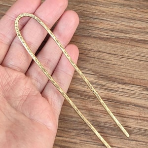 May include: A gold hair stick with a hammered texture. The stick is long and thin, and it is held in a hand.