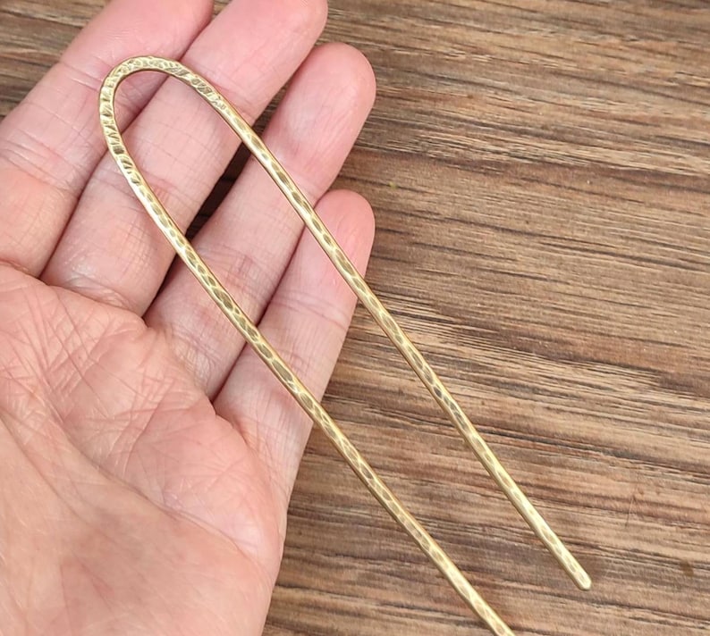 May include: A gold hair stick with a hammered texture. The stick is long and thin, and it is held in a hand.
