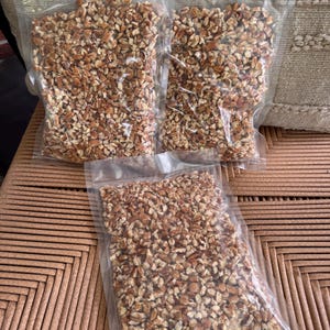 May include: Three clear, vacuum-sealed plastic bags filled with chopped brown and tan pecan pieces. The bags are rectangular and transparent, showcasing the texture and color of the nut fragments contained within each individual package.