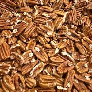 May include: A large collection of shelled pecan halves, showcasing their natural light to medium brown color, textured surface, and characteristic elongated, grooved shape. The nuts are piled together, filling the entire frame with their organic forms.