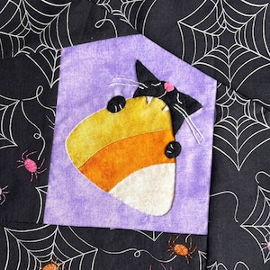 May include: A black cat with a candy corn in its mouth is nestled in a purple and black quilted fabric square. The fabric has a spiderweb pattern and the text "It's all mine" is on a metal tag above the square.