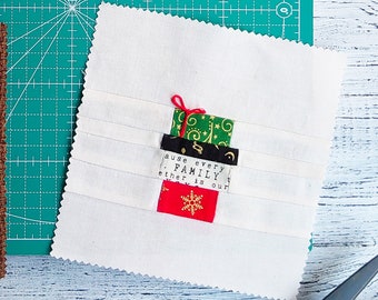 May include: Three fabric squares with appliqued gift box designs. One is on a brown background, two are on white. Sewing supplies, including pins, needles, and thread, are scattered around the squares on a green cutting mat.
