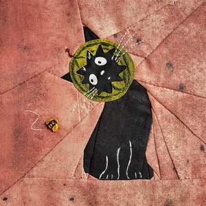 May include: A black cat with a green and yellow flower on its head is stitched onto a pink fabric background. The cat has white whiskers and a white outline. A small yellow bee is stitched on the fabric.