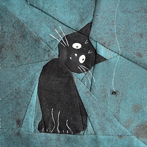 Könnte beinhalten: Eine schwarze Katze mit weißen Augen und Schnurrhaaren schaut nach rechts. Die Katze ist aus Stoff gefertigt und sitzt auf einem blauen und grauen Patchwork-Hintergrund. Ein weißes Band mit einer schwarzen Spinne am Ende hängt von der Oberseite des Bildes.