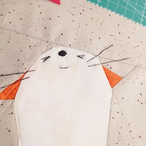 May include: A white fabric cat with orange ears and black embroidered whiskers and eyes. The cat is smiling and has a black nose. The cat is on a beige fabric background with brown dots.