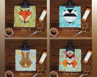 May include: Four fabric art pieces featuring animal faces: a fox, raccoon, rabbit, and squirrel. Each is clipped to a black clipboard, with text below. Sewing notions, including scissors and thread spools, are arranged around the pieces.