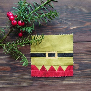 May include: A green fabric square with a black belt and a yellow buckle, representing an elf's outfit. The bottom of the square is red with a scalloped edge.