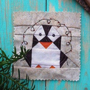 May include: A handmade patchwork penguin quilt block with black and white fabric and orange accents. The penguin is surrounded by a brown fabric border and a string of red and gold beads.
