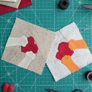 May include: Two fabric squares with a red, white, and orange geometric design. The squares are on a green cutting mat with a grid pattern.