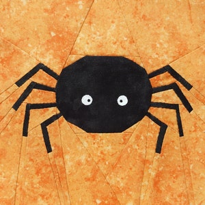 Könnte beinhalten: Eine schwarze Spinne mit weißen Augen auf orangefarbenem Hintergrund. Die Spinne ist aus Stoff gefertigt und hat ein einfaches Design.