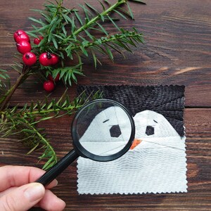 Puede incluir: Una lupa se utiliza para examinar una aplicación de tela con forma de pingüino. El pingüino es blanco con detalles negros y un pequeño pico naranja. La aplicación está sobre un fondo de tela gris oscuro.